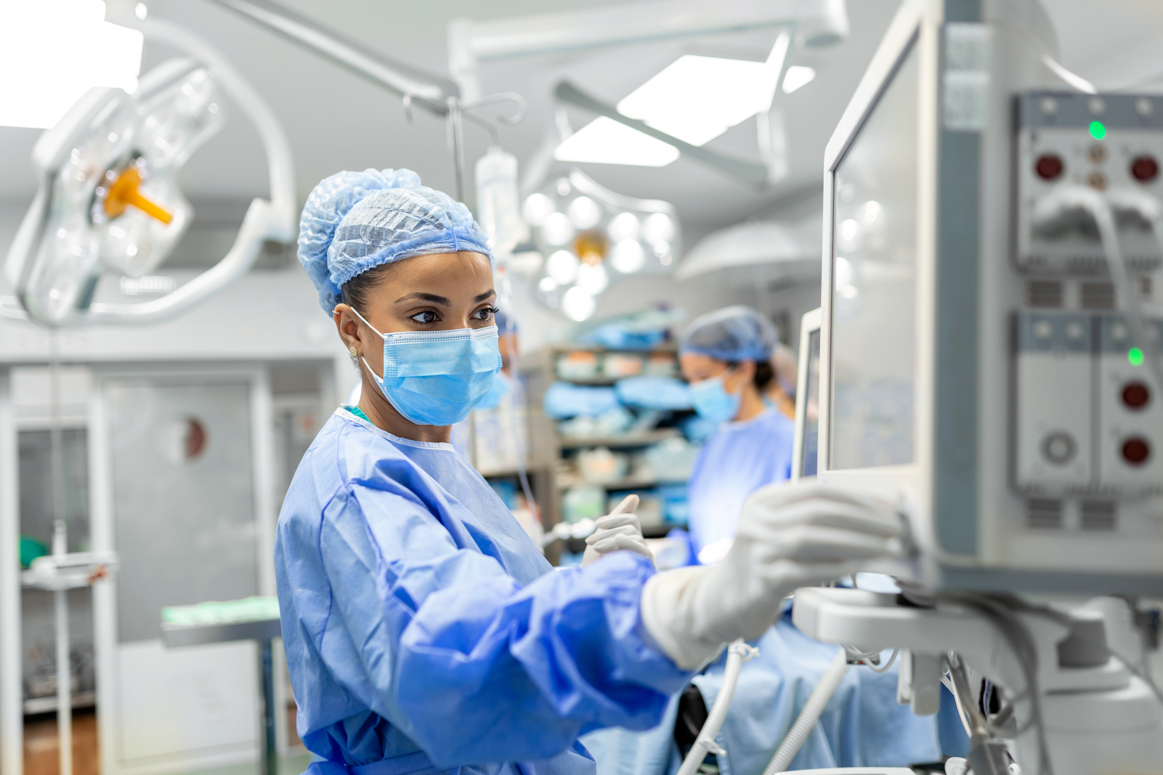 An anesthetist wearing blue scrubs and a mask and cap adjusts a machine in an operating room.