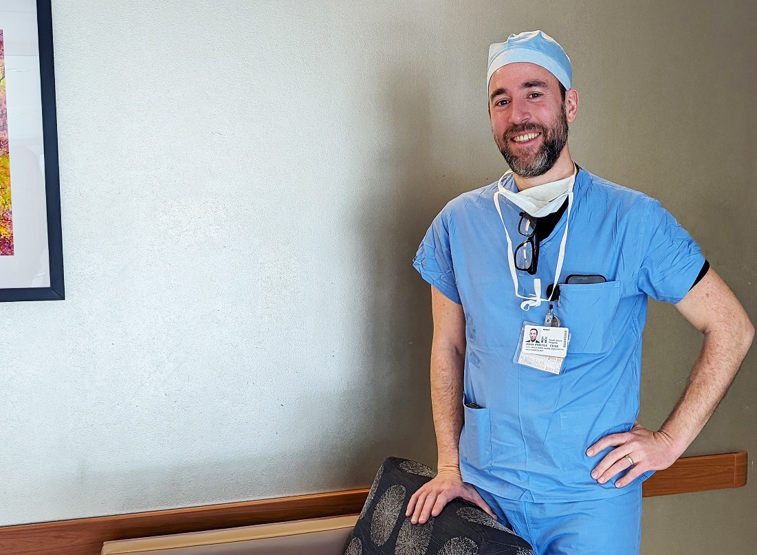 John Photos, CRNA poses for a photo wearing blue scrubs and a surgical cap