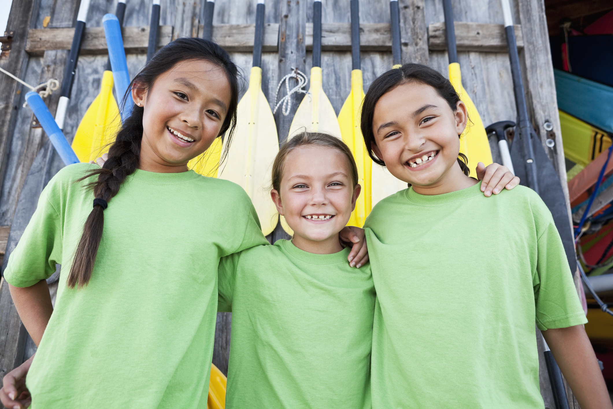 Three young girls wearing green shirts smile for a photo in front of oars at a pier of some kind. Wearing green for injury prevention day.