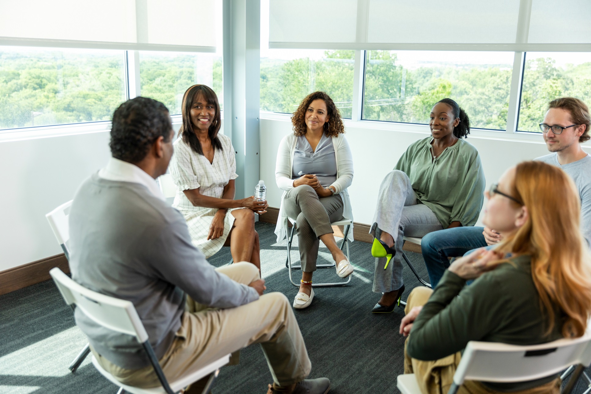 A mixed group of men and women sit in a circle of chairs in a support group setting.
