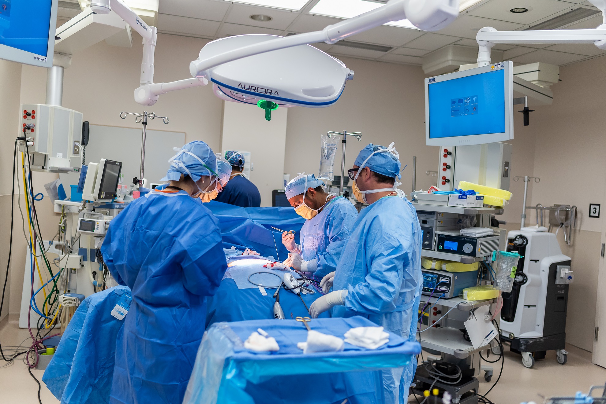 Three surgeons in blue scrubs, caps, and masks crowd around an operating table in an operating room at South Shore Hospital.