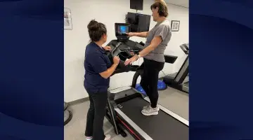 An exercise therapist points out functions on a treadmill while a class attendee stands on the treadmill listening.