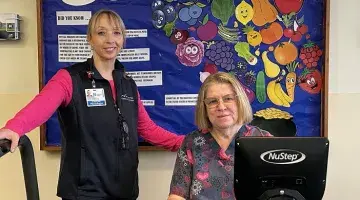 A female exercise physiologist stands next to a female cardiac rehab patient on an arm bike.