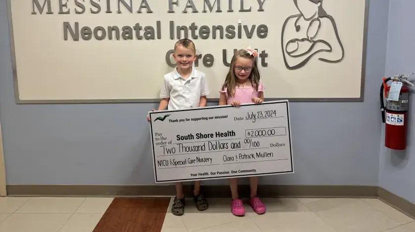 Patrick and Clara pose with a large check for $2,000 while standing in front of the NICU sign at South Shore Hospital