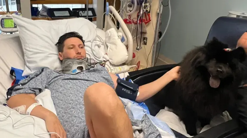 A man lying in a hospital bed with a black dog by his side