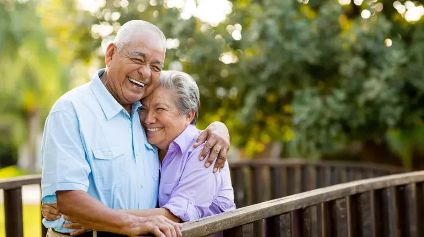 An older couple standing on a wooden bridge in nature laughs as they hug one another
