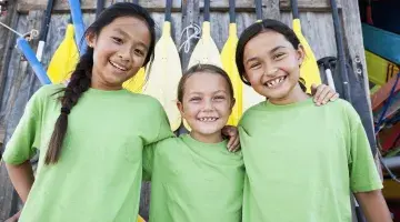 Three young girls wearing green shirts smile for a photo in front of oars at a pier of some kind. Wearing green for injury prevention day.