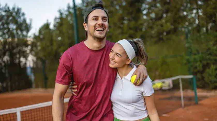 A young couple smiles as they hug after playing a game of tennis outdoors.