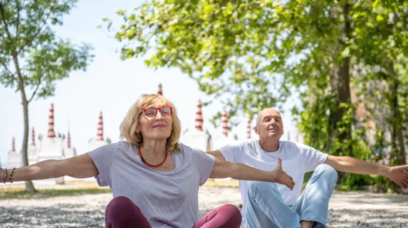 A man and woman sit barefoot on the ground with eyes closed and arms outstretched, seemingly deep-breathing or meditating.